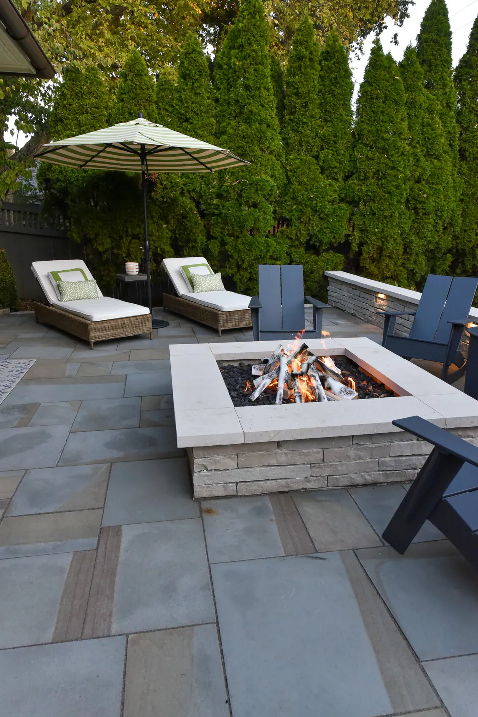 Square fire feature with chaise lounges, umbrella, evergreen privacy screen, and paver terrace